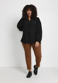 Bluză neagră cu mânecă lungă și decolteu în V, asortată cu pantaloni căptușiți maro. Modelul poartă pantofi negri cu o talpă texturată.