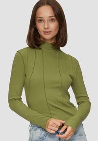 Haut vert à manches longues avec un col montant, présentant une texture côtelée et des détails de couture contrastants sur les côtés. Porté avec un jean en denim.