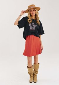 Př oversized černém tričku s textem "HOLD YOUR HORSES", oranžové sukni po kolena, béžových volných kozačkách a širokým hnědým kloboukem.