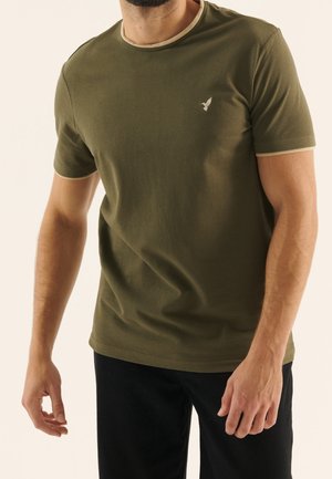 Mand iført olivengrøn kortærmet t-shirt med beige kant og broderet fuglelogo på brystet, kombineret med sorte bukser.