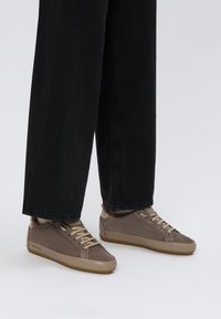 Baskets en suède marron avec des accents beiges, lacets plats et semelle intermédiaire texturée, associées à un pantalon noir ample.