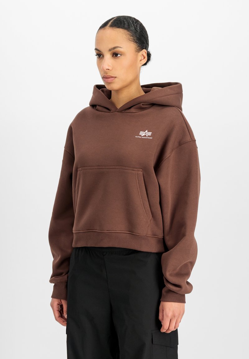 Γυναίκα που φοράει καφέ φούτερ Alpha Industries με μπροστινή τσέπη και μαύρο παντελόνι cargo, στέκεται μπροστά σε απλό λευκό φόντο.