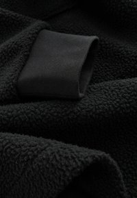 Sort fleece-stof med en blød, struktureret overflade. Har en glat, sort manchet i den ene kant, hvilket forbedrer designkontrasten.