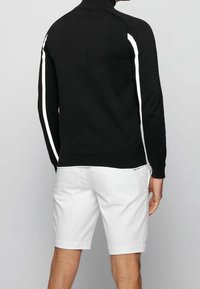 Svart stickad tröja med hög krage, med vita sidstripe; tillsammans med vita shorts som visar hud och en neutral bakgrund.