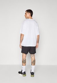 T-shirt blanche oversized, shorts de sport noirs avec accents blancs sur les côtés, complétés par des baskets noires et vertes et des chaussettes blanches.