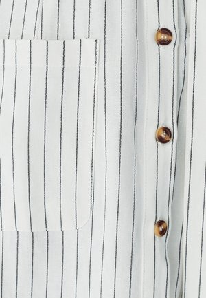 Bílá látková košile s černými vertikálními pruhy, která má levou náprsní kapsu a hnědé knoflíky vpředu. Hladká textura.