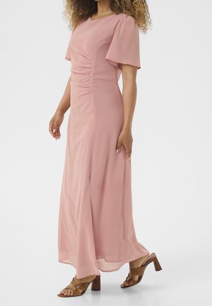Femme portant une longue robe rose clair avec des détails froncés et des manches volantes, associée à des sandales marron à talons.