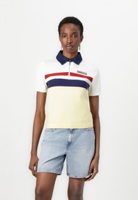 Polo t-shirt i hvid, gul og marineblå med lynlås i kraven og vandrette striber. Har et logo-patch på brystet. Kombineret med denim shorts.