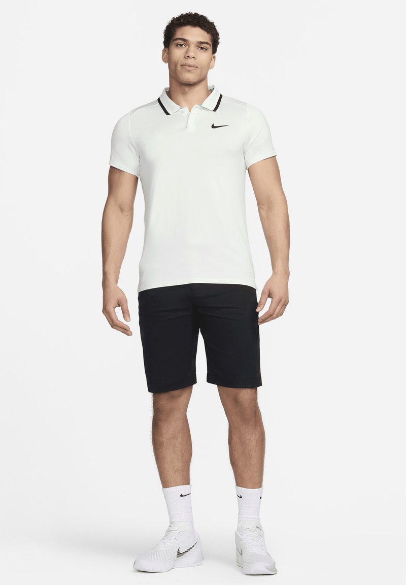 Camisa polo blanca con acentos de color negro en el cuello, logo en el lado izquierdo del pecho; combinada con pantalones cortos negros, calcetas blancas y zapatillas deportivas blancas.