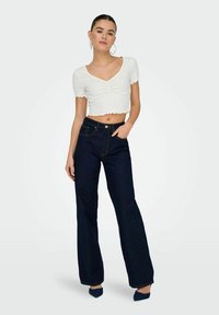 Witte cropped top met gegolfde randen, gecombineerd met donkerblauwe flare jeans. Model draagt puntige hakken, wat een strakke silhouet benadrukt.