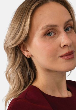 Frau mit hellblondem Haar, blauen Augen und natürlichem Make-up, die kleine goldene Ohrstecker und ein dunkelrotes Oberteil trägt, leicht seitlich blickend.