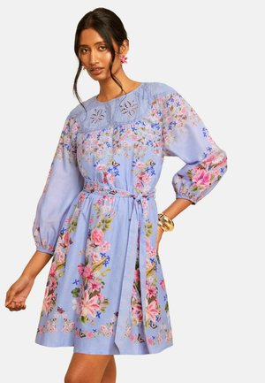 Femme portant une robe bleu clair à fleurs avec des manches bouffantes, une ceinture à la taille et des détails brodés découpés au niveau du décolleté, accessoirisée de bracelets dorés.