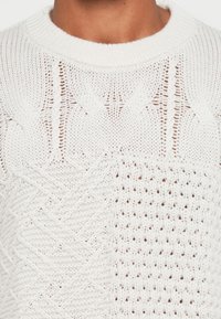 Fehér kötött pulóver, amely a kábel- és nyitott minták keverékét mutatja, texturális eltérésekkel és bordás kerek nyakkivágással.