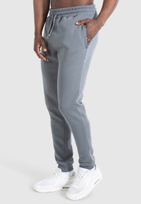 Pantalones de chándal grises con cinturilla elástica y cordón ajustable. Cuenta con bolsillos laterales y piernas estrechas. Combinados con zapatillas blancas.