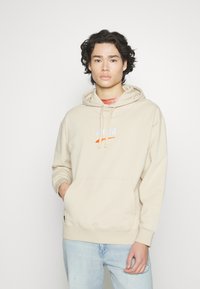 Beige hettegenser laget av mykt stoff, med en frontlomme, hettekord og hvit logo med oransje aksent på brystet. Lyseblå jeans.