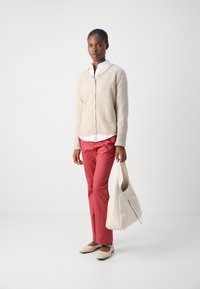Cardigan en maille beige, chemise blanche en dessous, pantalon rouge et chaussures plates beiges. Tient un sac hobo couleur crème. Design simple, texture douce.
