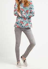 Camisa de mangas largas con fondo gris, que presenta vibrantes patrones rojos y verdes; combinada con jeans gris ajustados y zapatos gris de tipo slip-on.