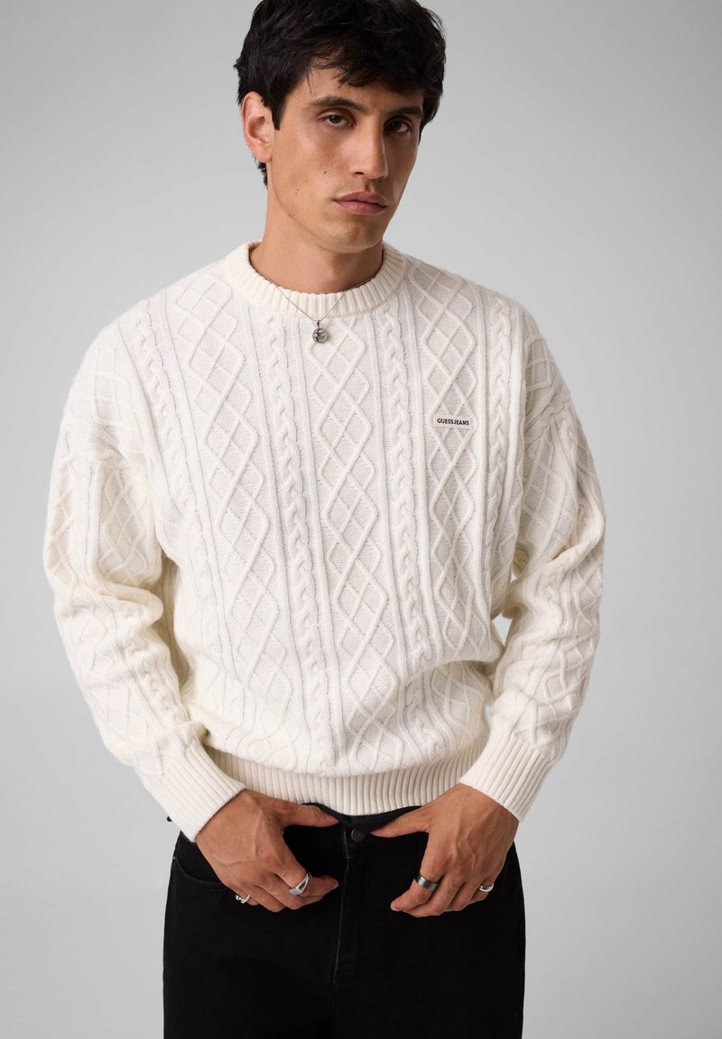 Homme portant un pull en maille torsadée blanc avec un petit logo sur la poitrine et un pantalon noir, debout sur un fond gris.