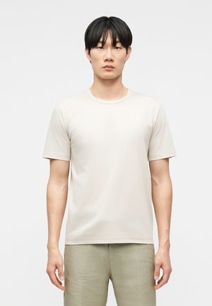 Jeune homme aux cheveux noirs et lisses portant un t-shirt beige clair à manches courtes uni et un pantalon de couleur claire, debout devant un fond blanc.