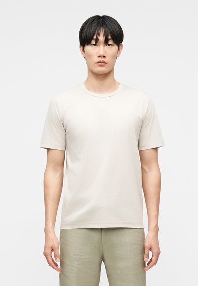 Junger Mann mit glattem schwarzen Haar, der ein schlichtes, hellbeiges kurzärmeliges T-Shirt und hellfarbene Hosen trägt, vor einem weißen Hintergrund stehend.