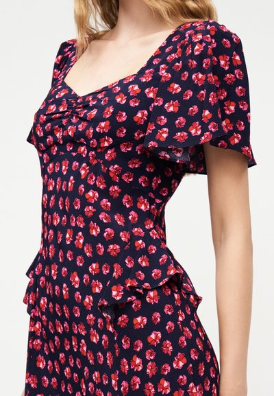 Γυναίκα που φοράει φόρεμα ναυτικού μπλε με κόκκινο και ροζ λουλουδάτο σχέδιο, με μανίκια που κυματίζουν και εφαρμοστό, ραμμένο μπούστο με λεπτομέρειες πεπλουμ.