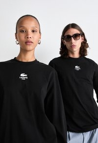 Zwei Models tragen schwarze Lacoste Paris Langarmshirts; eines mit Ohrringen, das andere mit Sonnenbrille und gestreiften Hosen, vor einem einfarbigen Hintergrund.