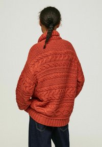 Ruosteenvärinen ylisuurikokoinen neulepaita, jossa yksityiskohtaiset palmikkokuviot, korkea kaulus ja resorireuna. Pehmeä rakenne ja rento istuvuus.