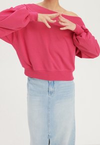 Felpa fucsia scollata con maniche arricciate, abbinata a una gonna di jeans azzurri con spacco frontale. Materiale in morbido cotone.