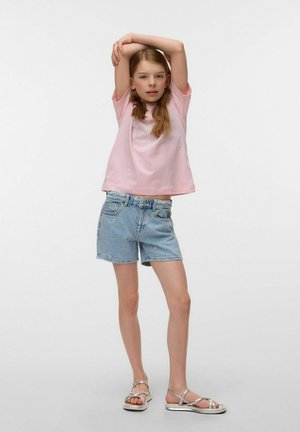 Ung pige står med armene løftet, iført en lyserød t-shirt, lyseblå denimshorts og sølvfarvede sandaler mod en ensfarvet baggrund.