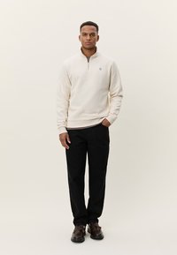 Μπεζ φούτερ με φερμουάρ, με ριμπ μανσέτες και τελείωμα, συνδυασμένο με μαύρο παντελόνι. Το σχέδιο διαθέτει ένα μικρό λογότυπο στο αριστερό στήθος, φορεμένο με σκούρα παπούτσια.