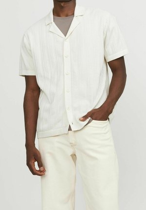 Homme portant une chemise boutonnée à manches courtes texturée crème par-dessus un t-shirt taupe avec un pantalon blanc cassé, main dans la poche, fond uni.