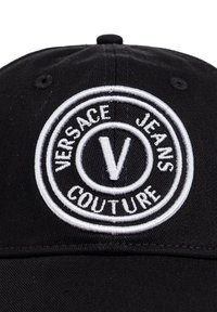 Casquette noire avec un logo rond brodé en blanc portant "VERSACE JEANS COUTURE" et un "V" proéminent au centre. Texture de tissu lisse.