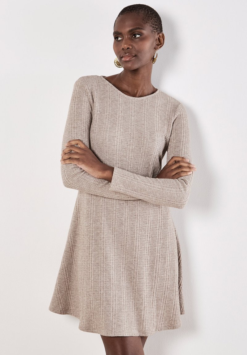 Vestido de punto de manga larga en beige claro con un patrón texturizado, ajuste entallado y falda acampanada. El modelo posa con los brazos cruzados.