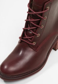 Gros plan d'une botte à talon haut en cuir bordeaux avec des lacets passés dans des œillets métalliques et des coutures détaillées sur le côté.