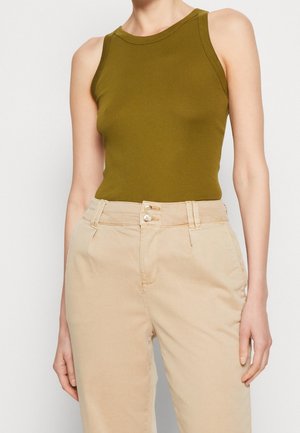 Person trägt ein olivgrünes ärmelloses Tanktop, das in beige hoch taillierte Hosen mit doppeltem Knopfverschluss gesteckt ist.
