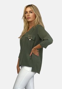 Oliwkowy top z dekoltem w serek i rękawami o długości trzech czwartych. Posiada przednią kieszeń ozdobioną łańcuszkiem i detalem w postaci zawieszki. Noszony z białymi spodniami.