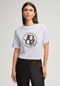 Witte katoenen T-shirt met een cirkelvormig logo in zwart en goud met textuurdetails. Korte mouwen en een relaxte pasvorm.