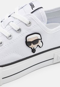 Sneaker bianco in tela con suola bianca e nera, lacci bianchi e un piccolo volto di cartone animato con occhiali da sole e cravatta sul lato.