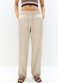 Kvinde iført beige drawstring brede bukser med hænderne i lommerne, parret med brune stroppsandaler og en cropped ærmeløs top.