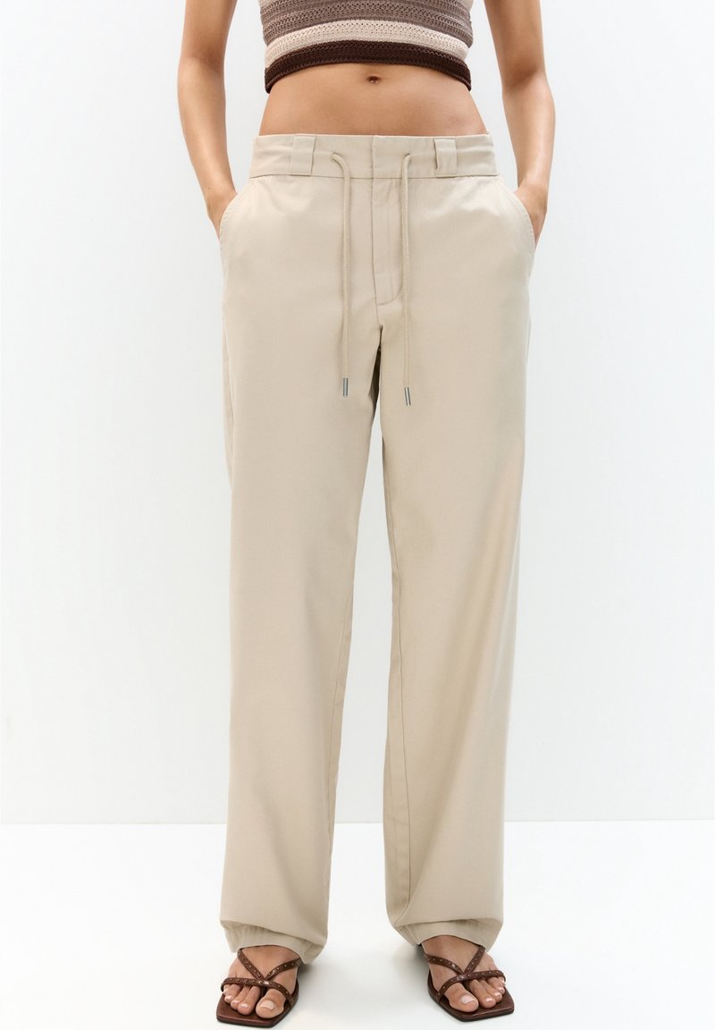 Kvinde iført beige drawstring brede bukser med hænderne i lommerne, parret med brune stroppsandaler og en cropped ærmeløs top.