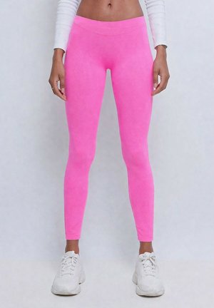 Femme portant des leggings rose vif, un top blanc court à manches longues et des baskets blanches épaisses, debout devant un fond clair uni.
