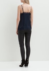 Kvinna i svart spetstopp med spetskant, svarta skinny jeans och svarta högklackade öppna ankelboots, stående mot en vit bakgrund.