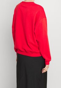 Czerwona, oversize'owa bluza z zaokrąglonym dekoltem, długimi rękawami i ściągaczami, w połączeniu z czarną spódnicą, widziana od tyłu.