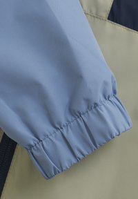 Helle blaue Jackenärmel mit gerafftem, elastischem Bund, der die glatte Stofftextur und ein kontrastierendes Panel in olivgrün zeigt.
