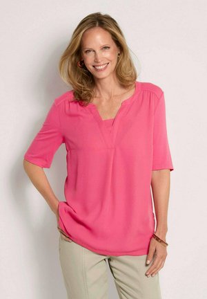 Femme souriante aux cheveux blonds mi-longs, portant un chemisier rose à manches courtes et un pantalon beige, debout avec une main sur la hanche.