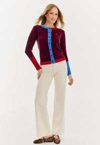 Burgundowa kardigan z niebieskimi guzikami i czerwonymi akcentami, z okrągłym dekoltem, fakturowanym materiałem, połączona z maślanymi spodniami i butami w leopardzie cętki.