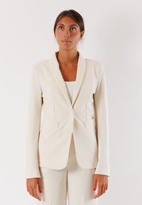 Blazer beige su misura con colletto classico, chiusura a un bottone e due tasche frontali. Presenta una texture liscia e una vestibilità su misura.