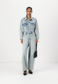 Lys blå denim jumpsuit med en tettsittende topp med to brystlommer, vide bukser og belte i midjen. Svart håndveske og sko.