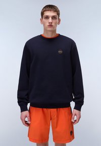 Marineblå genser med lange ermer, rund hals og liten sirkulær logo. Kombinert med klare oransje shorts med sidelommer.
