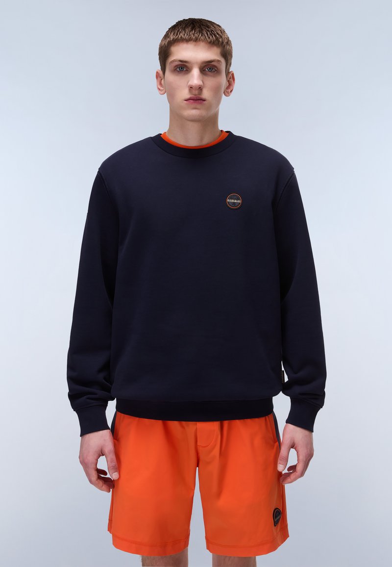 Marineblå genser med lange ermer, rund hals og liten sirkulær logo. Kombinert med klare oransje shorts med sidelommer.
